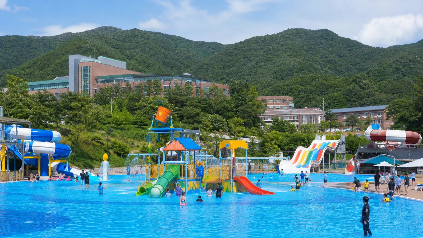 초록 산과 대학 건물을 배경으로 수영장과 물놀이 시설이 펼쳐진 괴산 여름 풍경